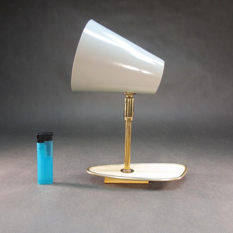 Pu&ograve; includere: Una lampada da scrivania vintage in ottone e bianco con un paralume a forma di cono. La lampada ha una base bianca con un bordo dorato.