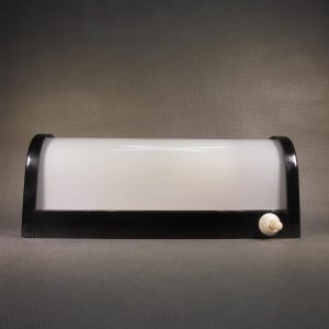 Może przedstawiać: Prostokątna lampa ścienna z zakrzywionym, półprzezroczystym białym szklanym kloszem w czarnej ramie. Podstawa jest czarna i posiada mały, okrągły, kremowy przełącznik przyciskowy umieszczony w dolnej prawej części oprawy.