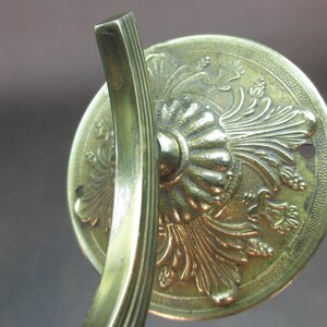 May include: A gold-toned metal wall mount with an ornate design featuring a stylized floral pattern. The mount has a curved arm and a circular base.
