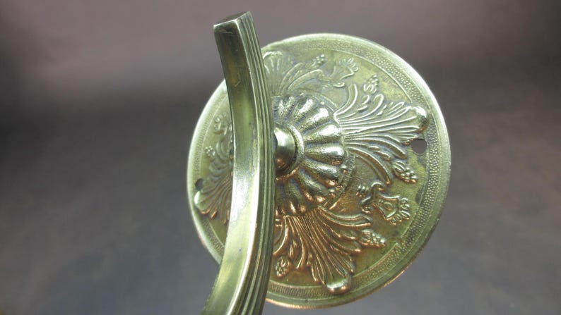 May include: A gold-toned metal wall mount with an ornate design featuring a stylized floral pattern. The mount has a curved arm and a circular base.