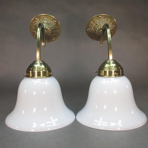 May include: Pair of brass wall sconces with white frosted glass shades. The sconces have ornate details and a curved arm.