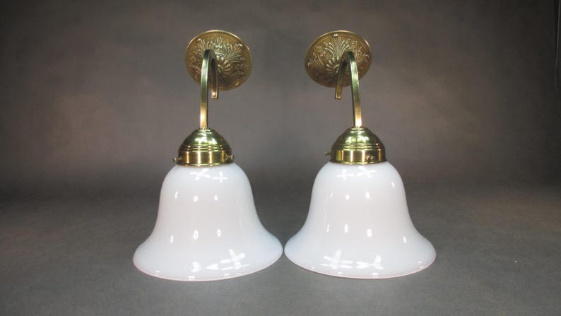 May include: Pair of brass wall sconces with white frosted glass shades. The sconces have ornate details and a curved arm.