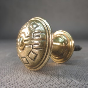 Może przedstawiać: Złoty metalowy gałka z ozdobnym, geometrycznym wzorem na górze i śrubą na dole.