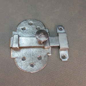 May include: A silver-colored metal latch with a circular base and rectangular arm. The latch has a knob in the center and several holes for mounting. The hardware is designed for securing doors or cabinets.