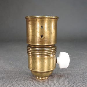 May include: A brass light bulb socket with a white porcelain switch. The socket has a threaded base and a small hole on the side.