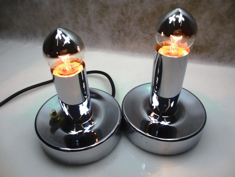 May include: Two chrome table lamps with round bases and clear glass bulbs. The bulbs are lit and casting a warm glow.