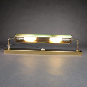 Könnte beinhalten: Eine Wandleuchte aus Messing mit zwei Glühbirnen. Die Leuchte hat eine lange, rechteckige Basis und einen zylindrischen Schirm mit einem matten Glasdiffusor.