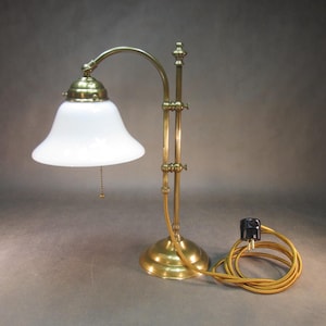 May include: A brass desk lamp with a white glass shade. The lamp has an adjustable arm and a round base. The cord is yellow and the lamp is plugged into a black outlet. The lamp is a classic design.