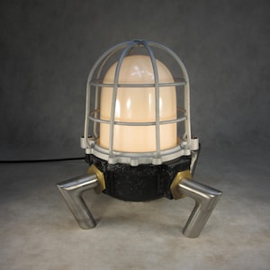 May include: A black and silver industrial style lamp with a cage-like shade and three curved legs. The lamp is turned on and the light is illuminating the room.