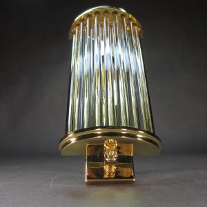 Op de afbeelding: Een goudkleurige wandlamp met een cilindrische, geribbelde glazen kap. De lamp heeft een rechthoekige basis en een gebogen bovenkant, allemaal in een glanzende gouden afwerking. De glazen staven zijn verticaal en creëren een textuureffect.