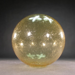 May include: A round, amber-colored glass object with a textured surface and small bubbles. The glass sphere has a slightly flattened base and reflects light, creating bright spots. The overall shape is a perfect sphere.
