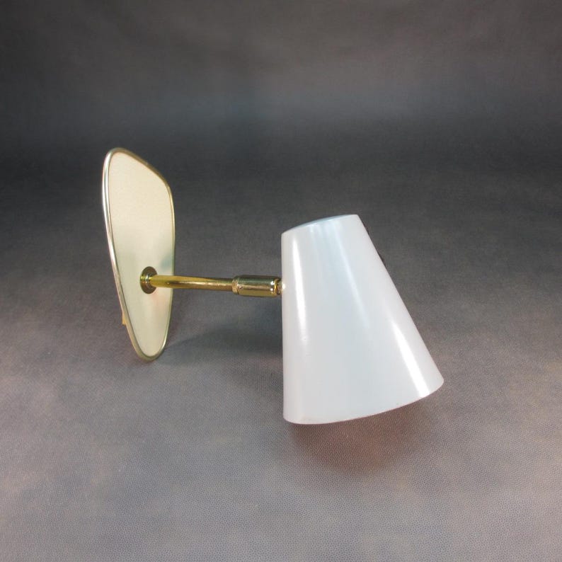 Pu&ograve; includere: Una lampada da parete in ottone con un paralume a forma di cono bianco. La lampada ha una piastra posteriore bianca curva.