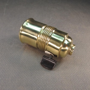 May include: A brass-colored light socket with a black switch. The socket has a cylindrical shape with ribbed detailing around the middle. The switch is a small, curved piece attached to the side.