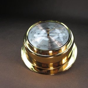 May include: A brass barometer with a white face and black needle. The face has markings for 700 to 800 millibars and the text "REGAL VENDERLICH SCHON" in black.