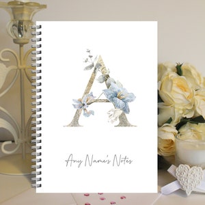 May include: A spiral bound notebook with a white cover featuring a gold glitter letter A decorated with blue and white flowers. The text "Any Name's Notes" is printed below the letter.
