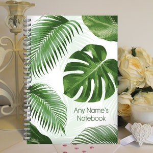May include: A white notebook with a spiral binding and a tropical leaf design. The notebook has the text "Any Name's Notebook" printed on the front.