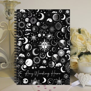 May include: Black and white spiral notebook featuring a celestial pattern of moons, stars, and suns. The text "Any Wording Here" is printed on the cover.