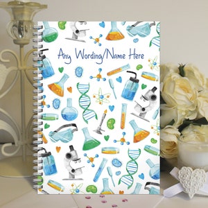 May include: A white spiral notebook with a colourful science theme. The cover features a repeating pattern of beakers, test tubes, microscopes, DNA strands, and other science-related imagery. The text "Any Wording/Name Here" is printed in blue on the cover.