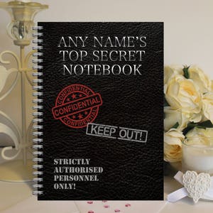 May include: Black notebook with a spiral binding. The cover has a faux leather texture and the text "ANY NAME'S TOP SECRET NOTEBOOK" in white. There are three red stamps that say "CONFIDENTIAL" and one red stamp that says "KEEP OUT!" The text "STRICTLY AUTHORISED PERSONNEL ONLY!" is printed in white below the stamps.