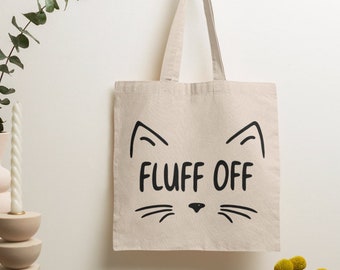 May include: A beige canvas tote bag featuring a black graphic print of a cat face outline with ears, whiskers, and a nose. The text "FLUFF OFF" is printed in black capital letters in the center of the cat face design on the front of the bag.
