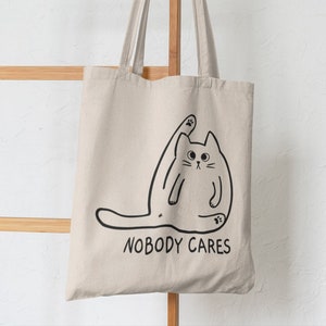 May include: A white canvas tote bag featuring a black line drawing of a cat stretching with the text "Nobody Cares" printed on the front.
