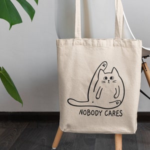 May include: A white canvas tote bag featuring a black line drawing of a cat with one paw raised in the air. The text "NOBODY CARES" is printed below the cat.
