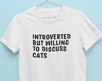May include: A white short-sleeved t-shirt hanging on a copper-coloured hanger. The front of the shirt features black text that reads: INTROVERTED BUT WILLING TO DISCUSS CATS. The shirt has a crew neckline and is made of a plain white fabric.
