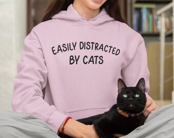 May include: A woman wearing a pink hoodie with the text "Easily Distracted By Cats" is holding a black cat in her lap.