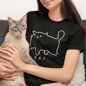 May include: A woman wearing a black t-shirt with a white outline of a cat and the word "cat" printed on it. She is holding a light brown cat in her arms.