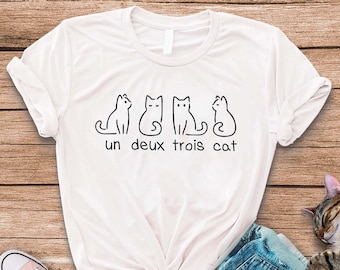 May include: White t-shirt featuring a graphic of three cats and the text "un deux trois cat". The shirt is laid out on a wooden surface with a pair of pink and white trainers and a pair of blue jeans. A brown tabby cat is sleeping on the jeans.