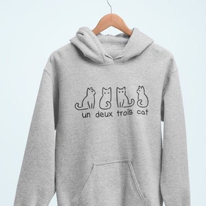 Op de afbeelding: Een grijs hoodie met een afbeelding van vier katten en de tekst "un deux trois cat".