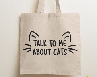May include: A beige canvas tote bag with two shoulder straps. The front of the bag features a black graphic print of cat ears and whiskers surrounding the text "TALK TO ME ABOUT CATS" in a bold, sans-serif font centered on the fabric surface.