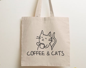 May include: A beige canvas tote bag featuring a black line drawing of a smiling cat holding a small mug. Below the cat illustration, the text COFFEE & CATS is printed in a simple black font. The bag has two long shoulder straps and a rectangular shape.