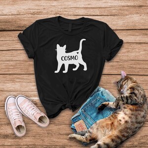 May include: Black t-shirt with a white silhouette of a cat with the word "COSMO" written on it. The cat has a small heart on its back.