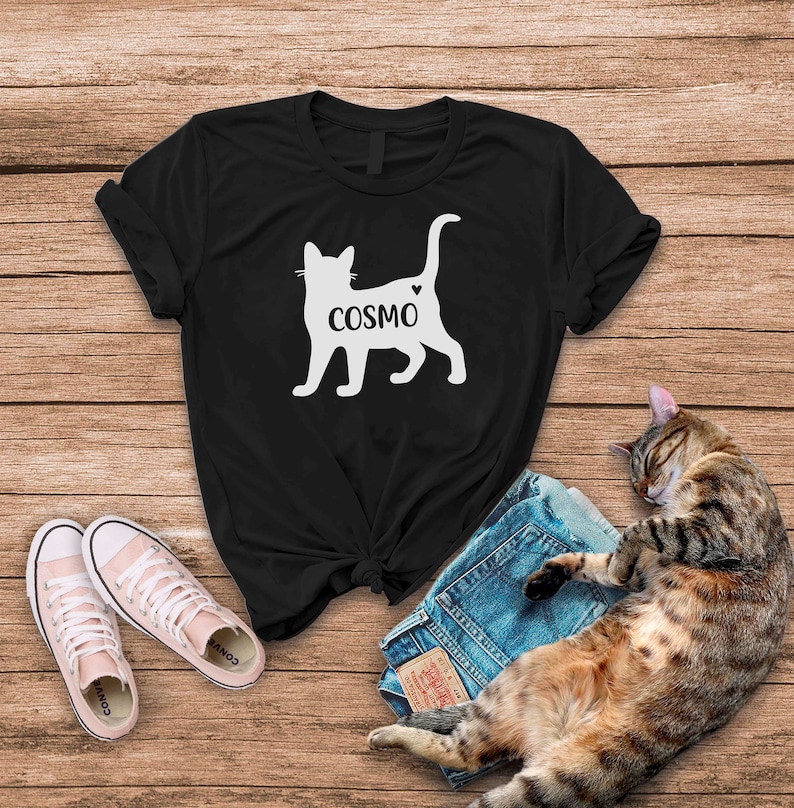 May include: Black t-shirt with a white silhouette of a cat with the word "COSMO" written on it. The cat has a small heart on its back.