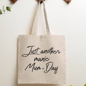May include: A beige canvas tote bag with black text that reads "Just another manic Mum-Day".