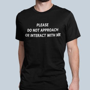 K&ouml;nnte beinhalten: Ein schwarzes T-Shirt mit dem Text "PLEASE DO NOT APPROACH OR INTERACT WITH ME" in wei&szlig; gedruckt.