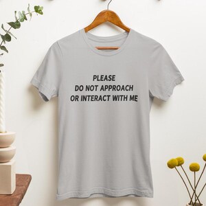 K&ouml;nnte beinhalten: Ein graues T-Shirt mit dem Text "PLEASE DO NOT APPROACH OR INTERACT WITH ME" in Schwarz gedruckt.