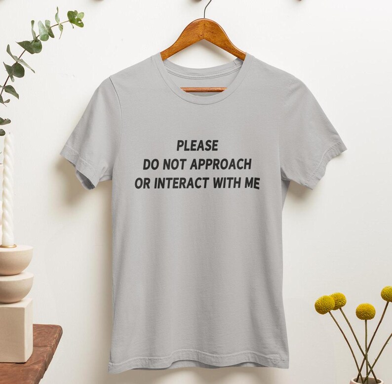 K&ouml;nnte beinhalten: Ein graues T-Shirt mit dem Text "PLEASE DO NOT APPROACH OR INTERACT WITH ME" in Schwarz gedruckt.