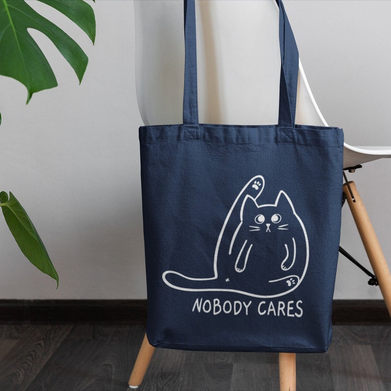 May include: A navy blue tote bag featuring a white outline of a cat with its paw raised in the air. The text "NOBODY CARES" is printed below the cat.