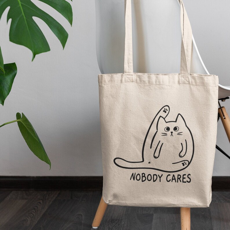May include: A white canvas tote bag featuring a black line drawing of a cat with one paw raised in the air. The text "NOBODY CARES" is printed below the cat.