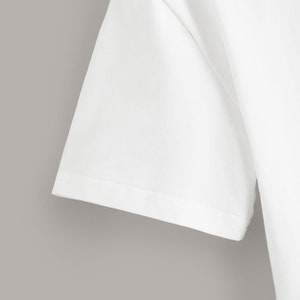 May include: Close-up of the white cotton fabric of a short-sleeved t-shirt. The fabric is smooth and has a subtle texture. The seam is visible along the edge of the sleeve.