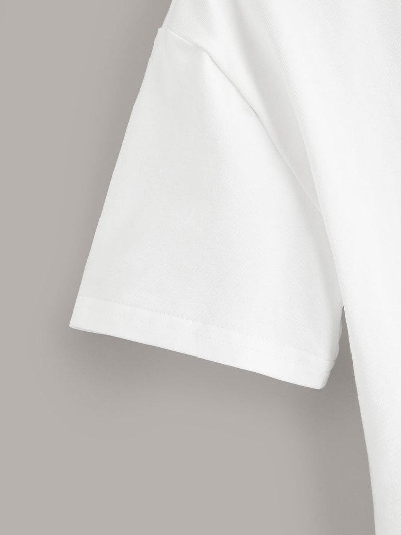 May include: Close-up of the white cotton fabric of a short-sleeved t-shirt. The fabric is smooth and has a subtle texture. The seam is visible along the edge of the sleeve.