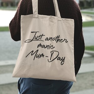 May include: A beige tote bag with black text that reads "Just another manic Mum-Day".