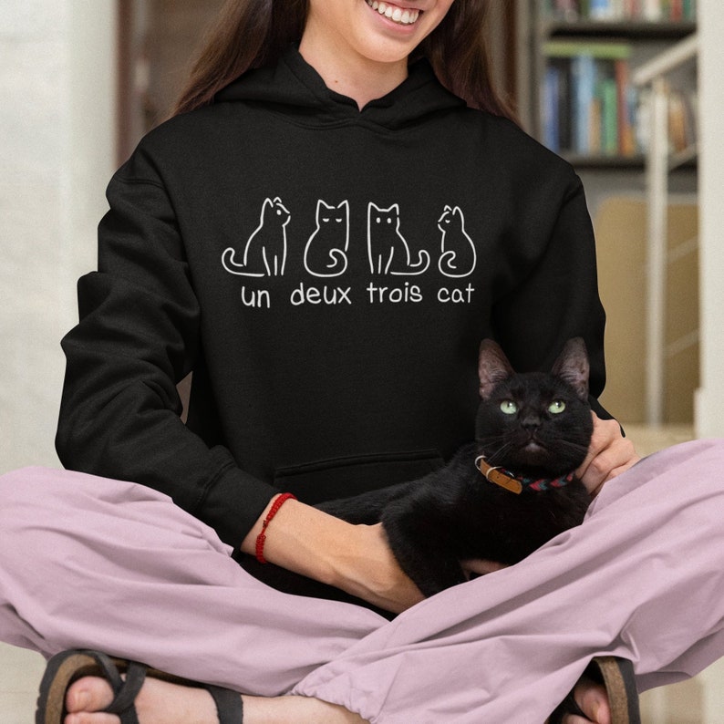 Op de afbeelding: Een zwarte hoodie met een witte afbeelding van drie katten en de tekst "un deux trois cat". Een persoon draagt de hoodie en houdt een zwarte kat vast.