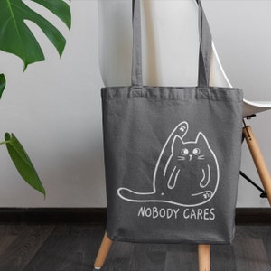 May include: A grey canvas tote bag featuring a white outline of a cat with its paw raised and the text "Nobody Cares".