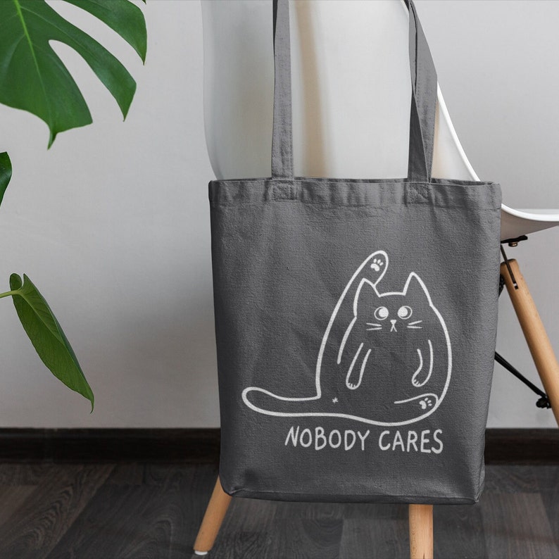 May include: A grey canvas tote bag featuring a white outline of a cat with its paw raised and the text "Nobody Cares".
