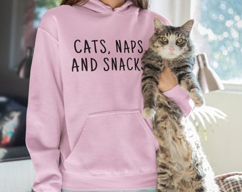 May include: A pink hoodie with the text "Cats, Naps and Snacks" printed on the front. A person is holding a tabby cat in their arms.