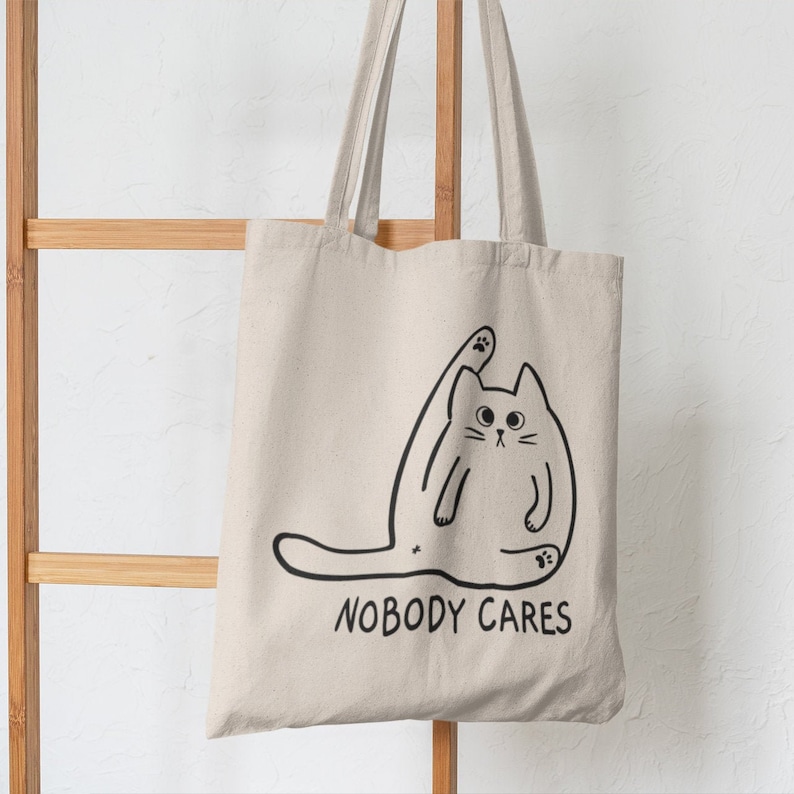 May include: A white canvas tote bag featuring a black line drawing of a cat stretching with the text "Nobody Cares" printed on the front.