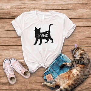 May include: White t-shirt with a black silhouette of a cat with the name "COSMO" printed on it. A brown tabby cat is sleeping on a pair of blue jeans.